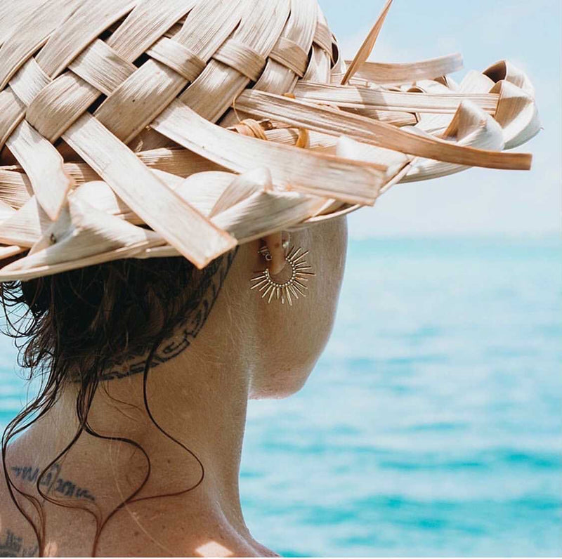 spiky yellow gold and diamond earrings on woman with straw hat on in front of the ocean