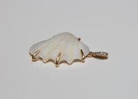 A scallop-shaped white shell pendant with gold-toned claw shaped prongs, and a small pavé-set diamond bail, shown on a plain light gray background.