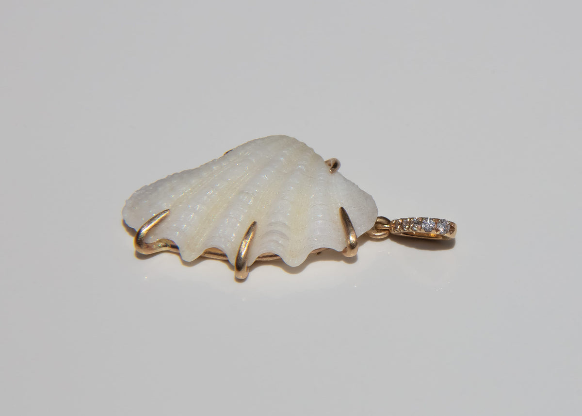 A scallop-shaped white shell pendant with gold-toned claw shaped prongs, and a small pavé-set diamond bail, shown on a plain light gray background.