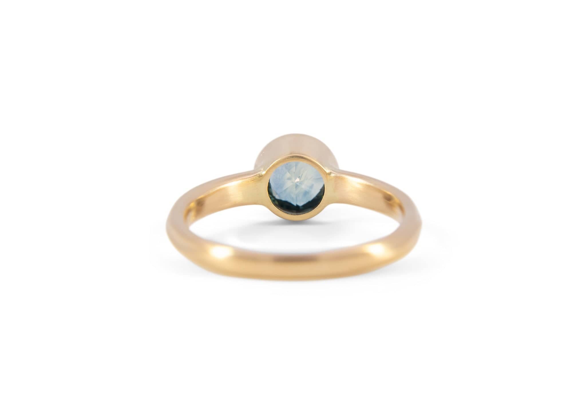back of a gold solitaire ring with a teal montana sapphire against a white background