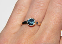 Gold solitaire ring with a teal montana sapphire on a hand against a white background