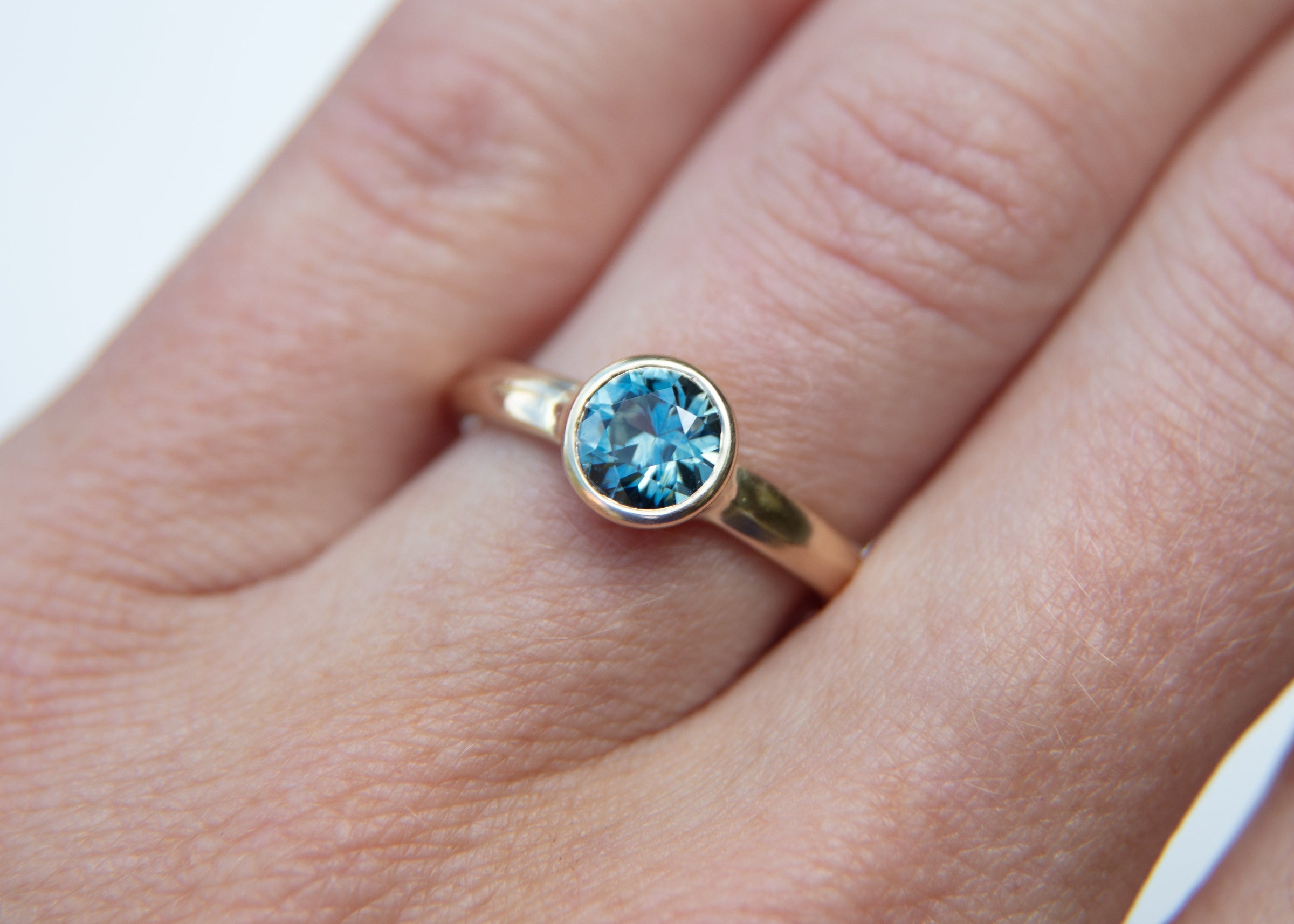 Gold solitaire ring with a teal montana sapphire on a hand against a white background